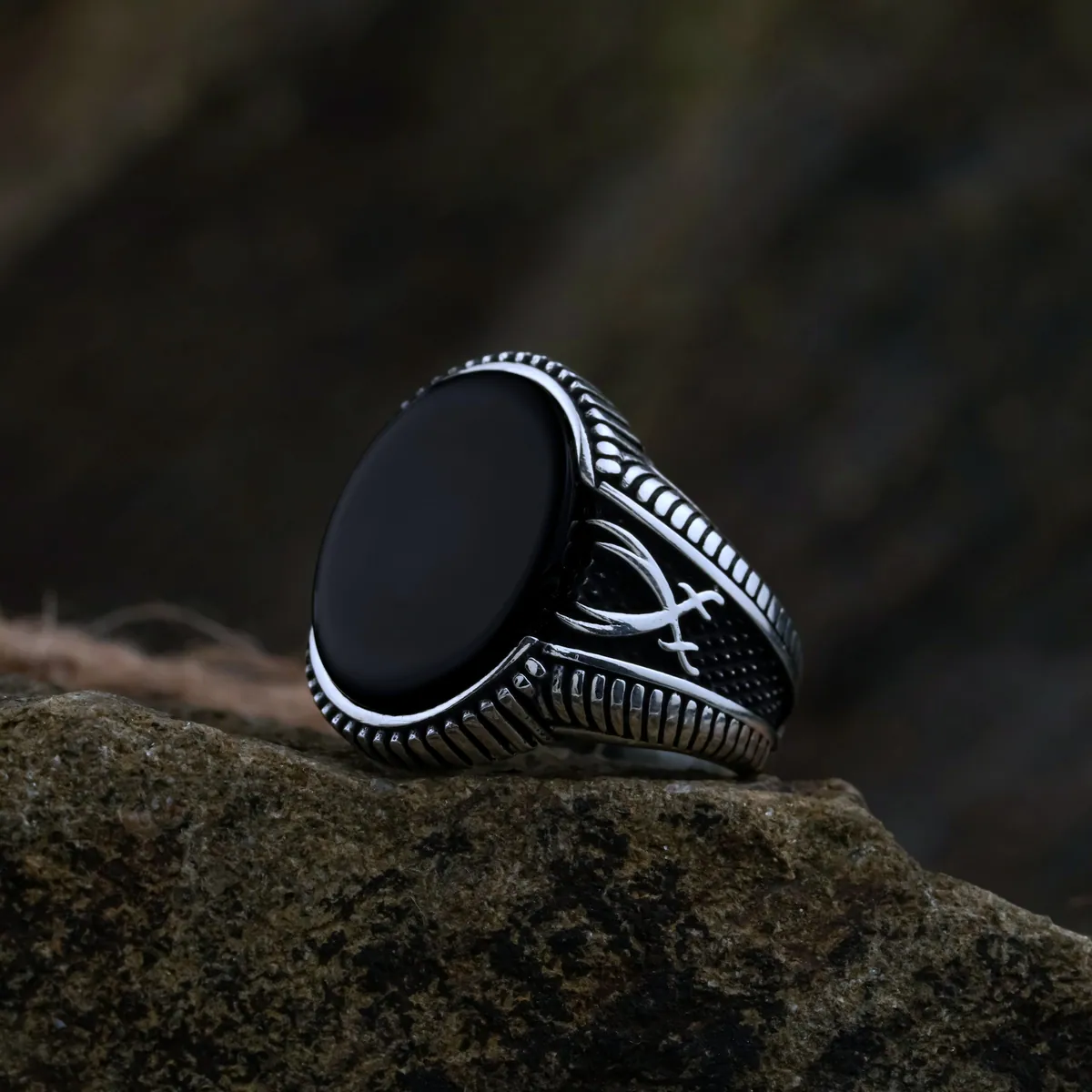 A silver signet ring with a black oval face and engraved shoulders, on dark rock