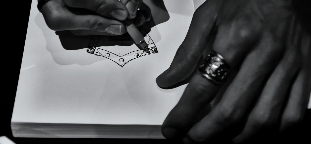 Black and white photo of hands drawing a design in a sketchbook, a heavy silver ring on one finger